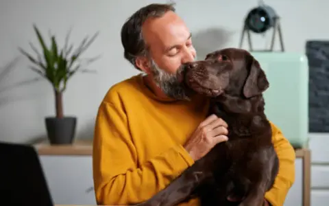 Hombre con barba besa a su perro chocolate. "No puedo seguir teniendo a mi perro, ¿qué hago?" se pregunta, abrazándolo.
