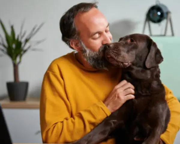 Hombre con barba besa a su perro chocolate. "No puedo seguir teniendo a mi perro, ¿qué hago?" se pregunta, abrazándolo.